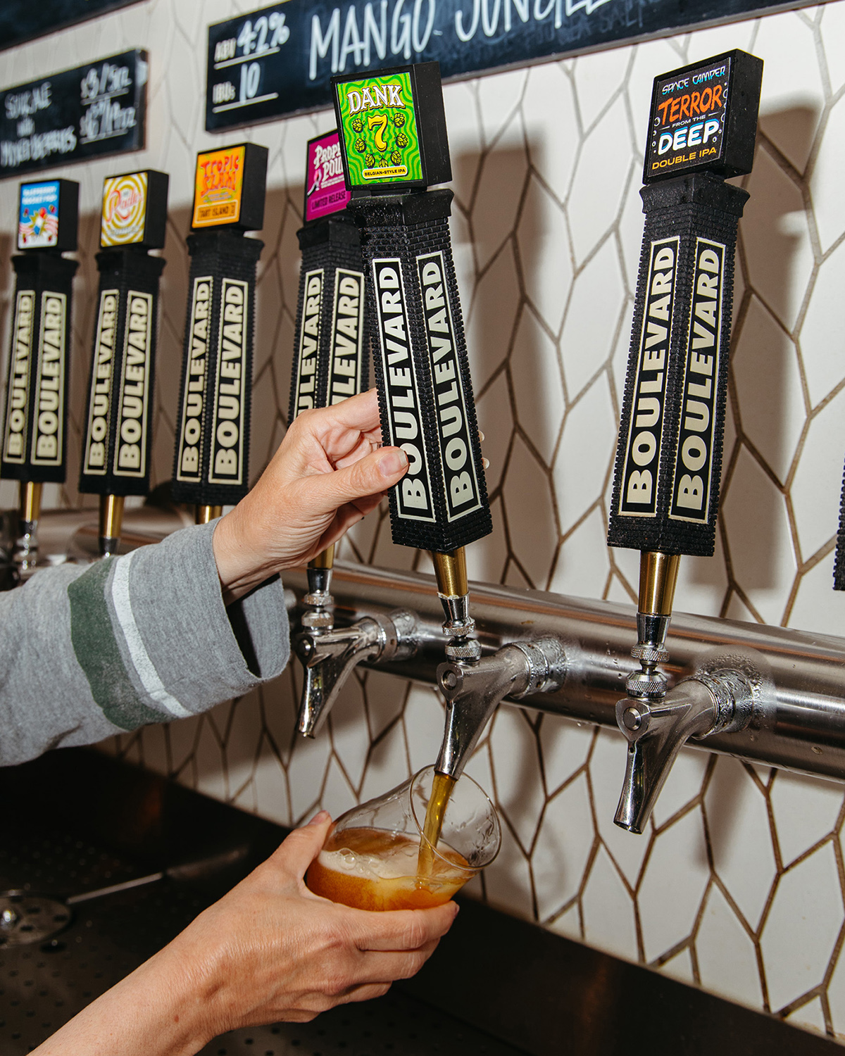 A person is filling a pint glass with beer from a tap labeled "Boulevard." The backdrop has tiled walls, and other taps with different labels are visible. The hand of the person pouring beer is the focal point of the image.