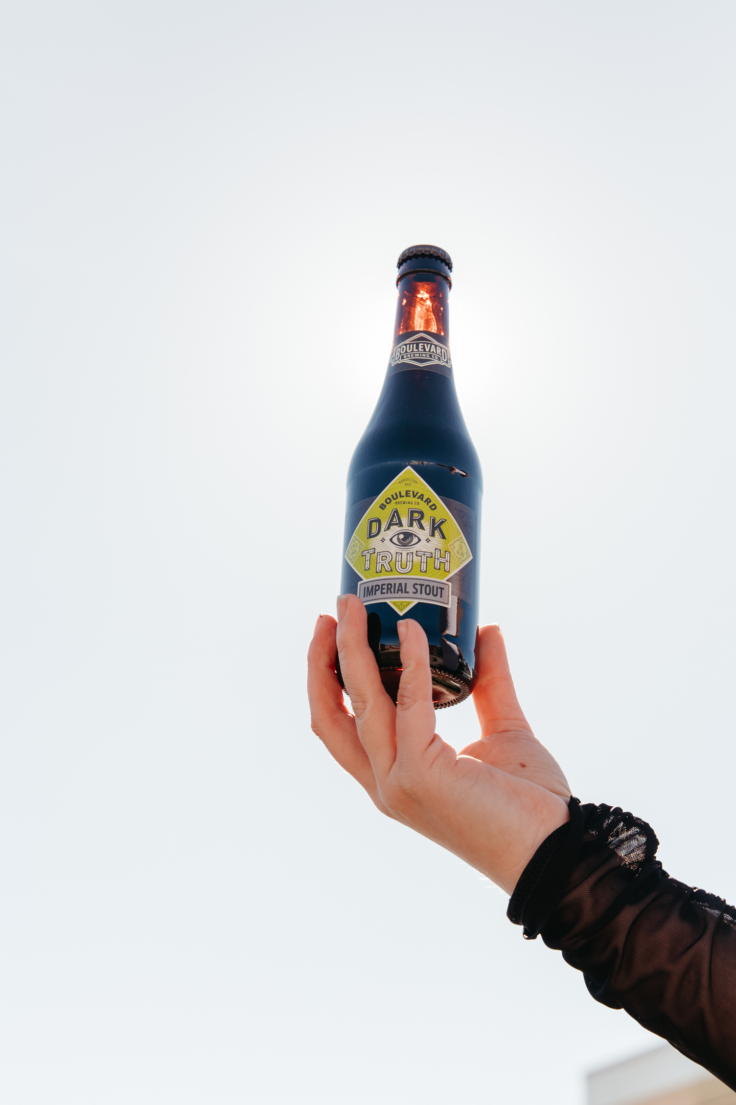 A hand holding a bottle of Boulevard Dark Truth Imperial Stout beer against a bright, clear sky. The bottle features a yellow and black label with the brand and beer name prominently displayed. The person is wearing a dark, sheer sleeve.