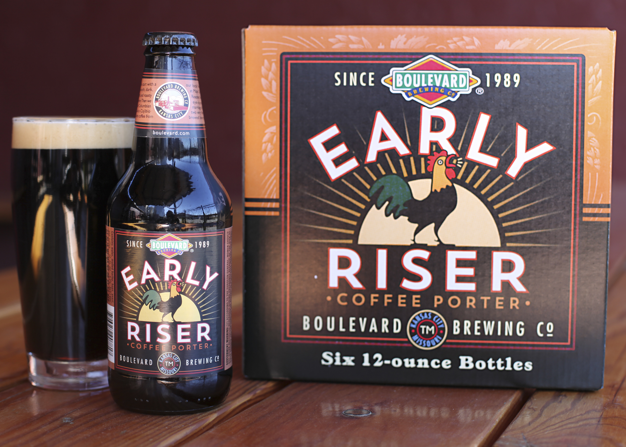 A bottle and six-pack of Boulevard Brewing Co.'s Early Riser Coffee Porter are displayed on a wooden table. The bottle is next to a filled glass showcasing the dark beer. The label features a rooster and vintage-style design.