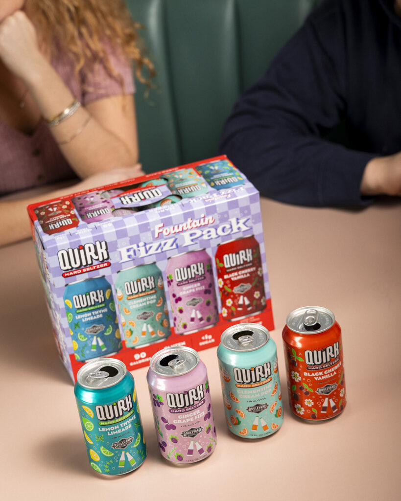 A colorful display of Quirk hard seltzer cans and a variety pack box, featuring four different flavors. The box and cans are placed on a table, with parts of two people, one in a pink top and another in a navy top, partially visible in the background.