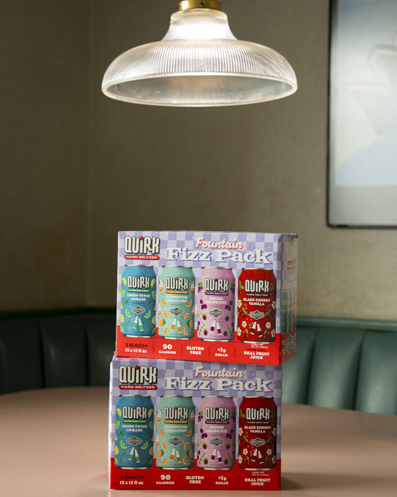 Two packs of Quirk Hard Seltzer's "Fountain Fizz Pack" are stacked on a table beneath a hanging light. Each pack contains four flavors: Lemon Thyme Limeade, Clementine Cream Pop, Ginger Grape Fizz, and Black Cherry Vanilla. A cushioned bench and artwork are in the background.