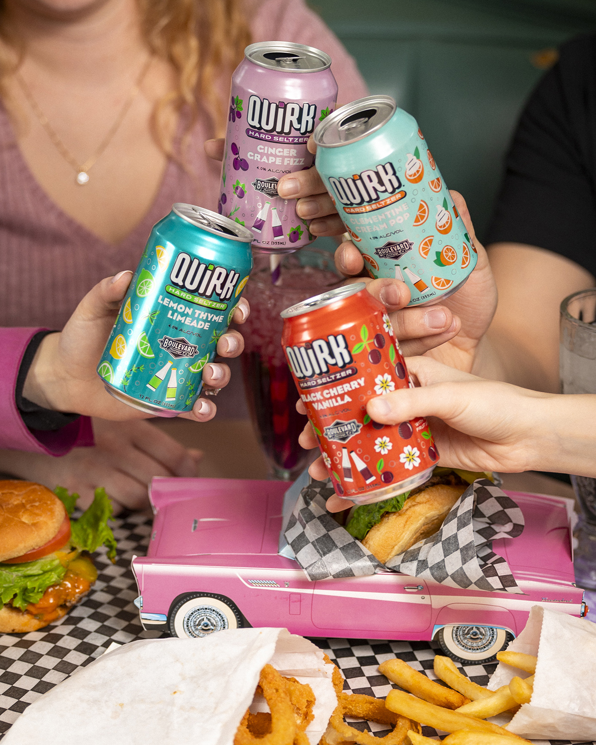 Four people are toasting with cans of Quirk Spiked & Sparkling seltzer, each holding a different flavor. Below, there's a pink vintage car tray holding a burger and fries. The background includes part of a table with more food and drinks.