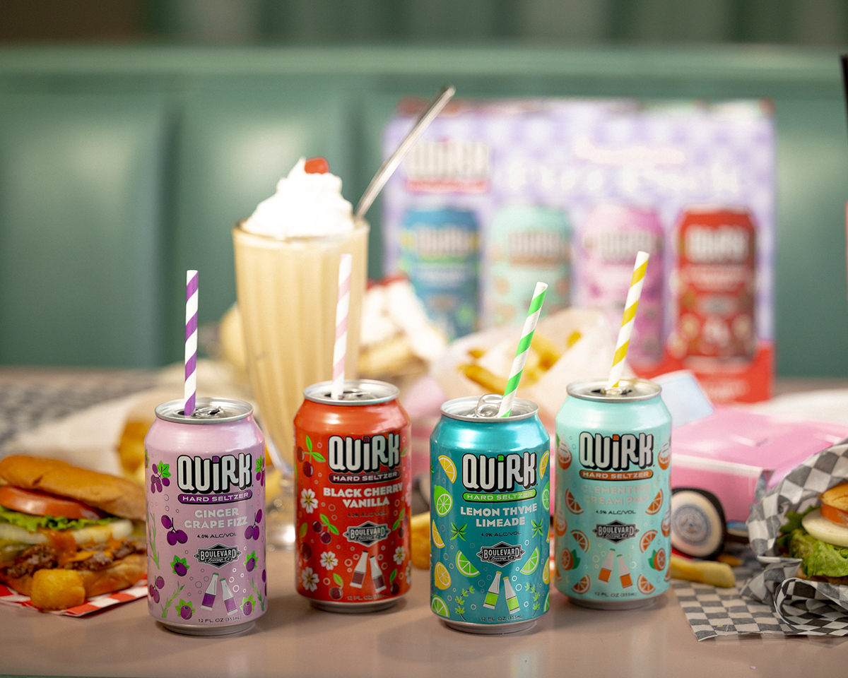 Four colorful cans of Quirk Hard Seltzer in flavors Ginger Grapefruit, Black Cherry Vanilla, Lemon Thyme Limeade, and Strawberry Lemon Basil are displayed on a retro-style diner table. Behind them are a milkshake, a hamburger, fries, and a box with Quirk branding.