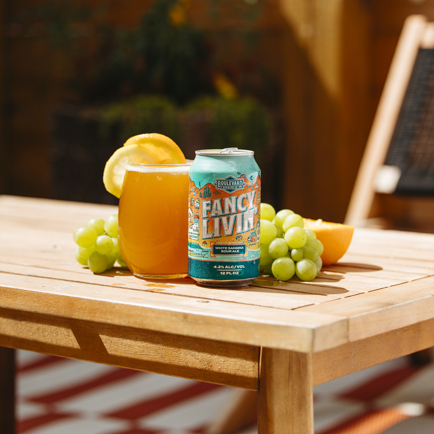 A can of Boulevard Fancy Livin' beer is placed on a wooden table next to a glass of beer garnished with an orange slice. Fresh grapes and an orange wedge surround the glass and can. The background features outdoor furniture and plants.