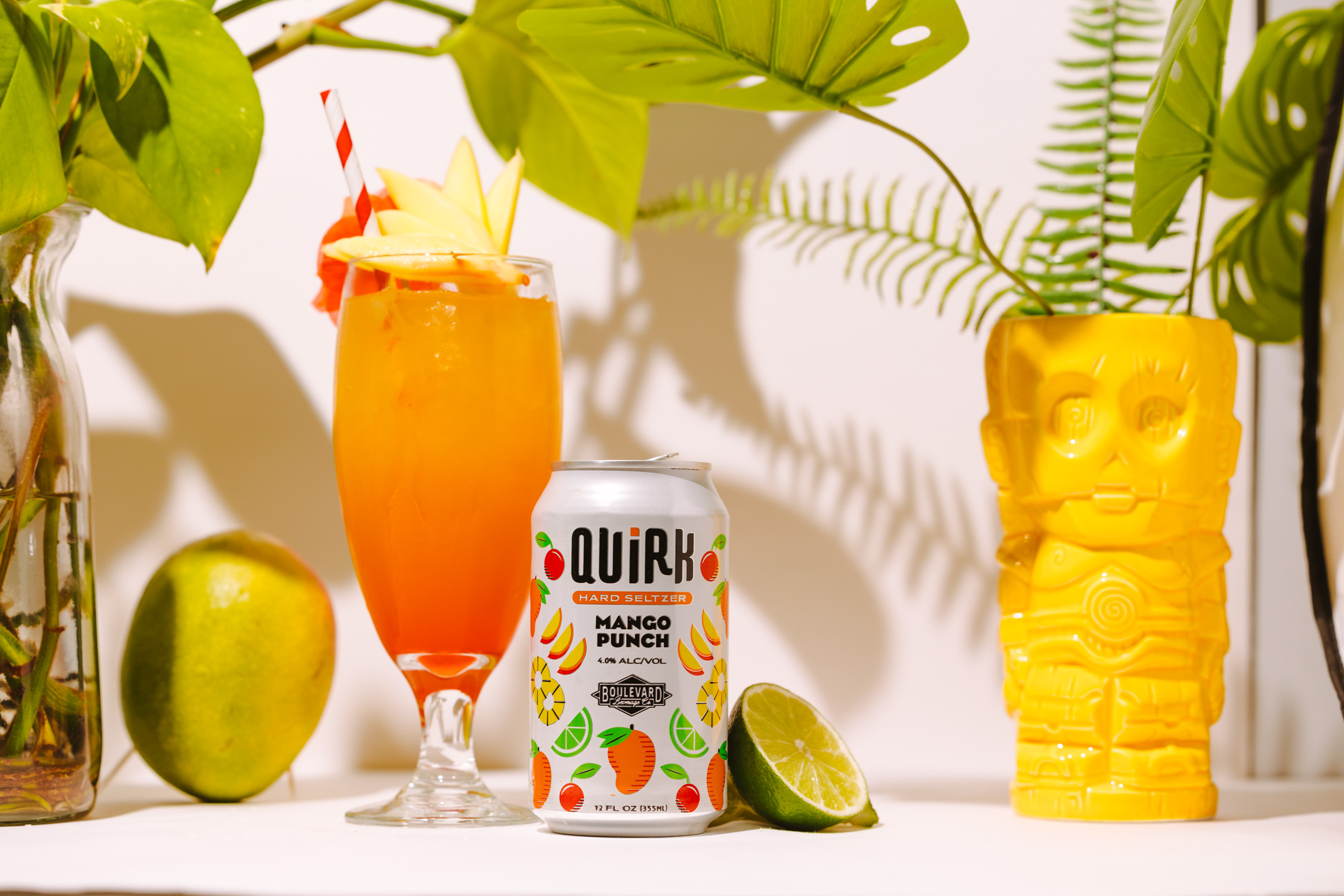 A tropical scene featuring a cocktail glass with a colorful drink and fruit garnish, a can of Quirk Mango Punch, fresh mango, lime wedge, and a yellow tiki mug. Background includes large green leaves and a fern.