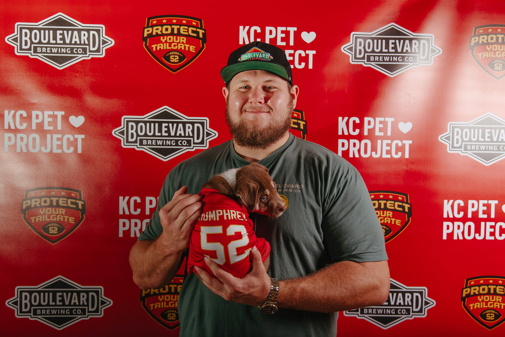 A man in a green shirt and black cap is holding a small brown puppy dressed in a tiny red jersey with "Humphrey 52" on the back. They are standing in front of a red backdrop featuring logos from Boulevard Brewing Co., Protect Your Tailgate, and KC Pet Project, clearly excited about the event.