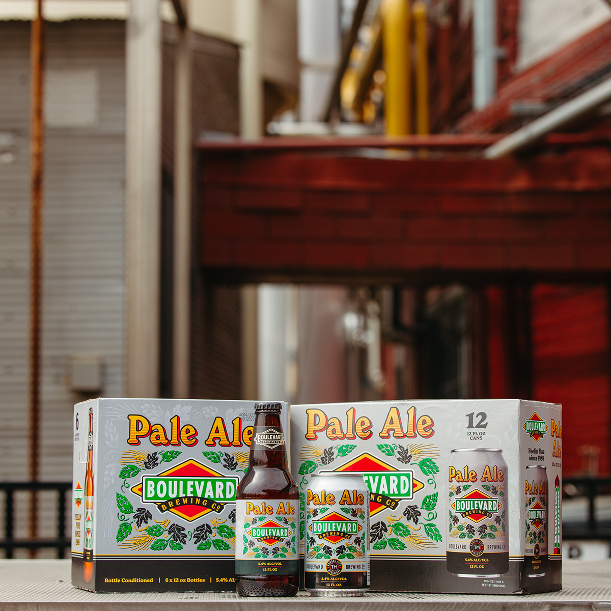 A case of Boulevard Brewing Co. Pale Ale beer is displayed outdoors on a metallic surface. In front of the box is a single bottle and can of Pale Ale. The building walls and pipes create an urban industrial backdrop.