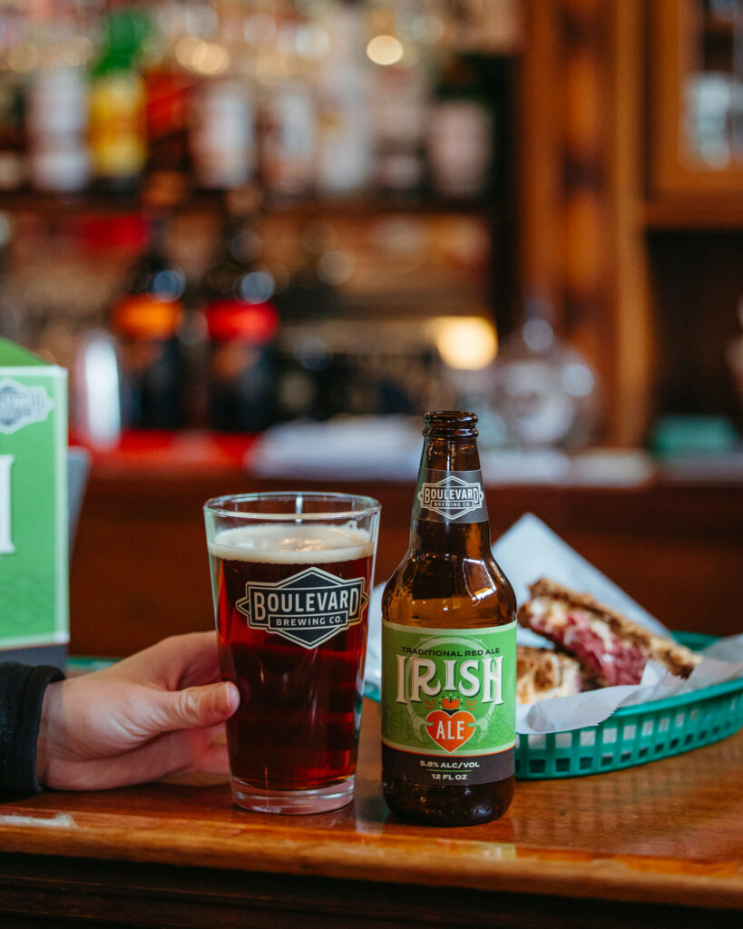 A person holding a glass of Boulevard Irish Ale next to a bottle of Boulevard Irish Ale on a bar counter. In the background, there are blurred bottles of liquor and a basket with food, possibly a sandwich, partially visible.