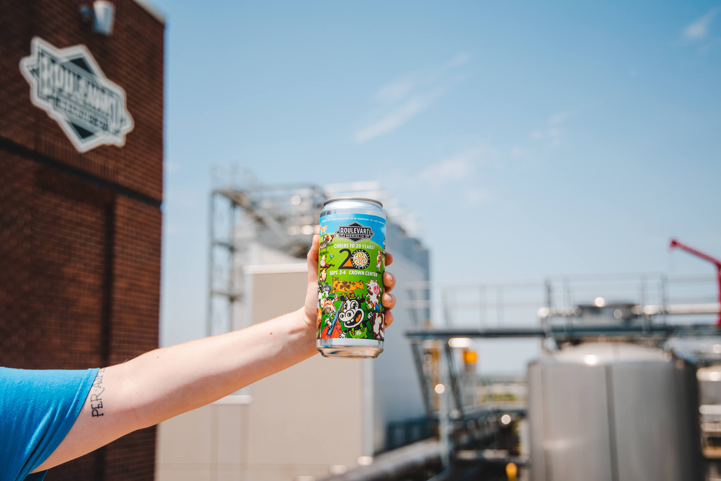 A person wearing a blue shirt holds up a colorful can of beer outdoors, with an industrial brewing facility in the background. The can has vibrant artwork and text that tells you all about the brew. The sky is clear and blue, suggesting a sunny day.