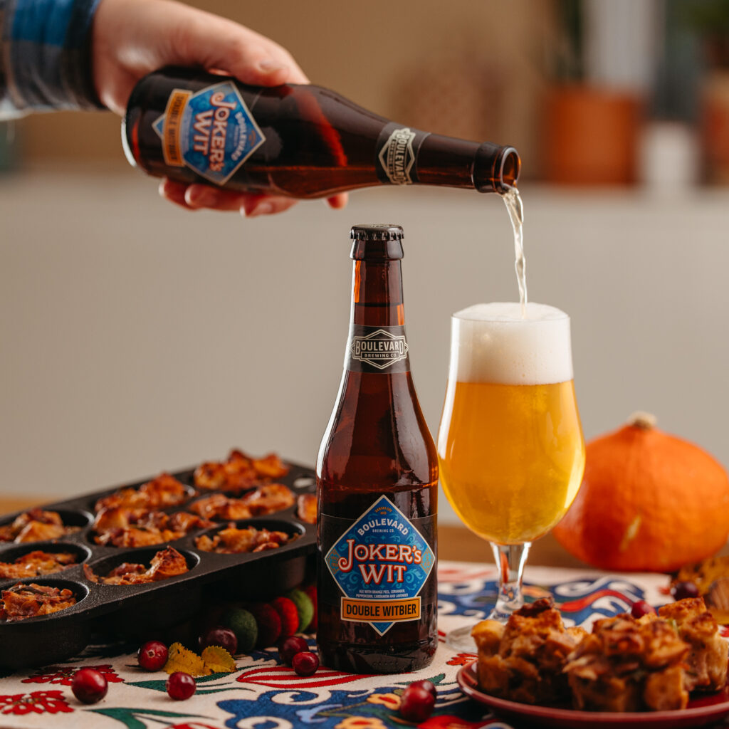 A hand pours Joker's Wit beer from a bottle into a glass. Another Joker's Wit bottle is on the table next to a tray of baked goods and a small pumpkin. The table is covered with a colorful cloth and scattered with a few cranberries.