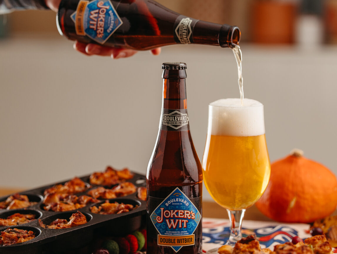 A hand pours Joker's Wit beer from a bottle into a glass. Another Joker's Wit bottle is on the table next to a tray of baked goods and a small pumpkin. The table is covered with a colorful cloth and scattered with a few cranberries.