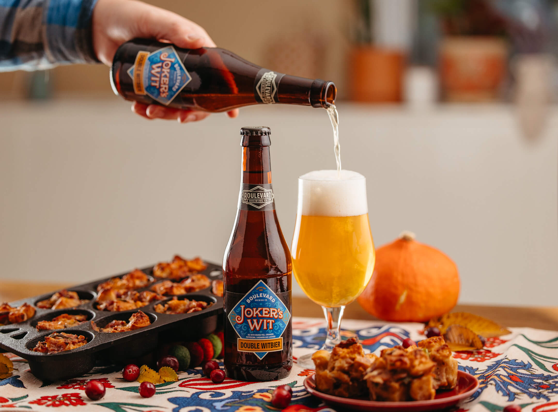 A person is pouring a bottle of Joker's Wit Double Witbier into a glass of beer. In the foreground, there is another full bottle of Joker's Wit, a plate of snacks, and a muffin tray with baked items. A small pumpkin can be seen in the background.