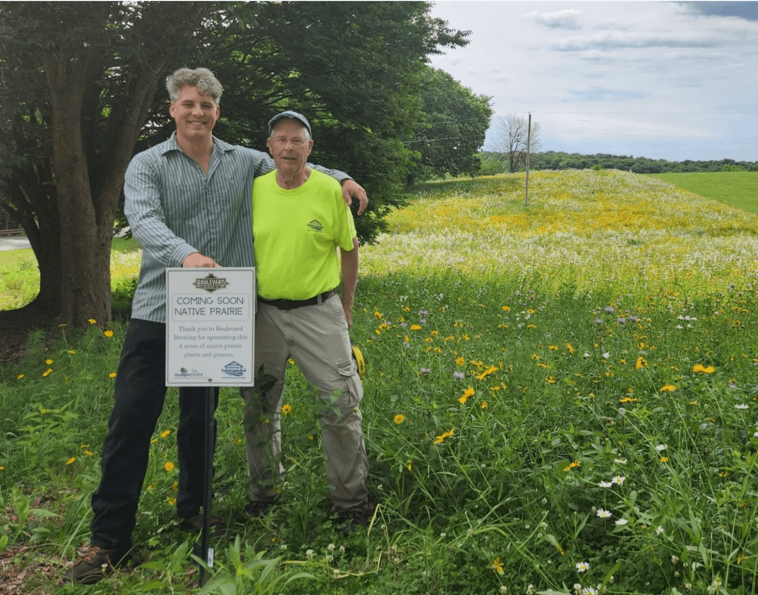 Two men stand in a grassy field filled with wildflowers, smiling at the camera. The man on the left is in a striped shirt and the man on the right in a neon green T-shirt. They both hold a sign that reads "COMING SOON NATIVE PRAIRIE" with additional text below.