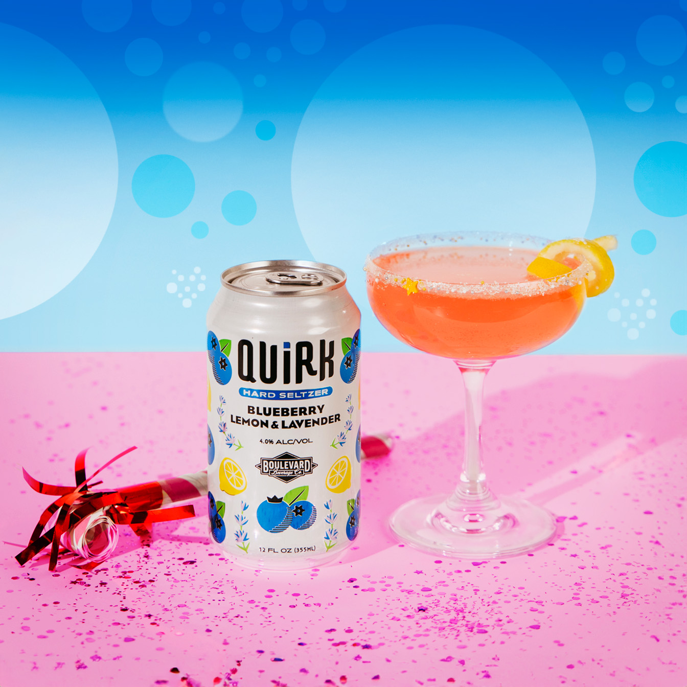 A can of Quirk Blueberry Lemon & Lavender hard seltzer is next to a cocktail glass filled with a pink drink, garnished with a lemon twist. The background has blue circular patterns and the surface is pink with confetti and a red party horn.