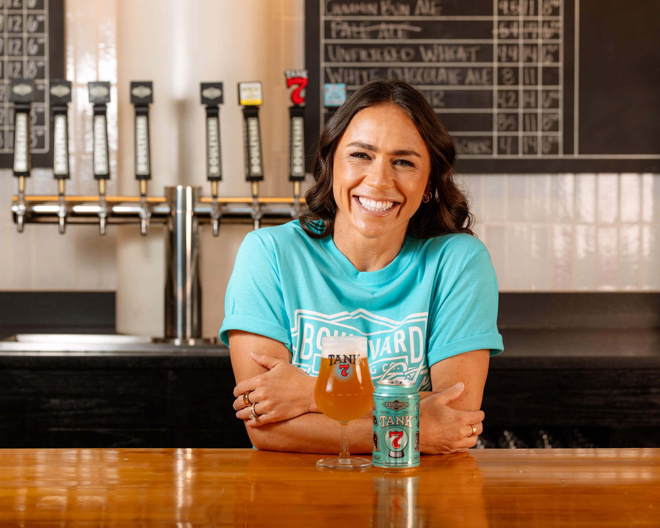 A person with long dark hair and a big smile wears a turquoise T-shirt and sits at a wooden bar. A beer glass and a branded beer can are on the bar in front of them, and beer taps and a chalkboard with drink options are visible in the background.