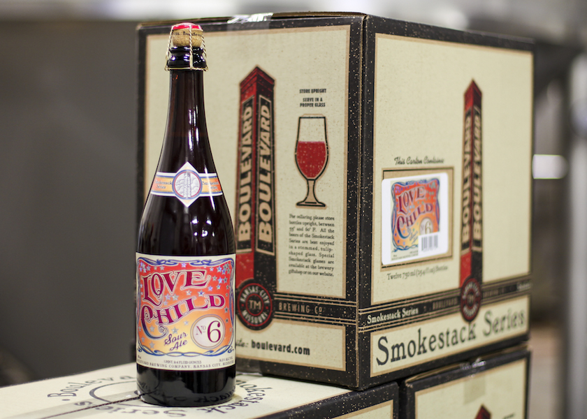 A bottle of "Love Child No. 6" beer stands in front of a box that features the same label design. The label is colorful with red and yellow accents. The box is part of the Smokestack Series from Boulevard Brewing Co.