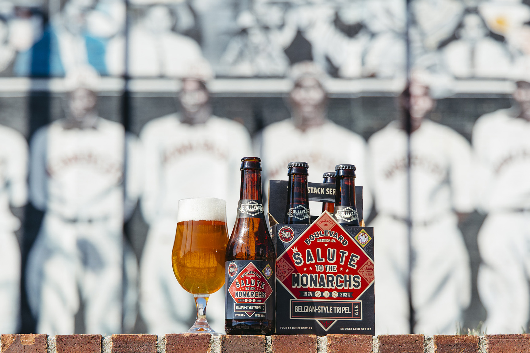 A six-pack of Boulevard Brewing Company's "Salute to the Monarchs" Belgian-Style Tripel beer and a poured glass of beer are displayed outdoors. The background features a black-and-white mural of vintage baseball players.