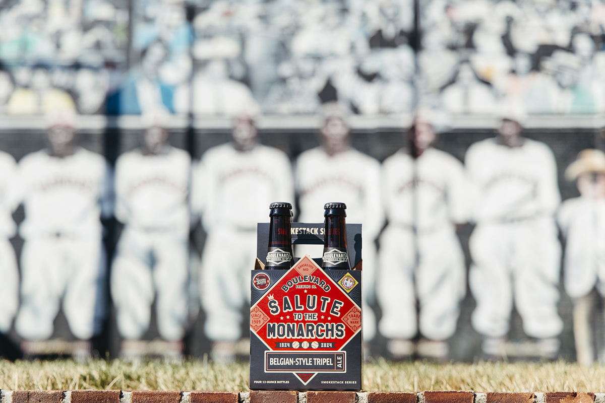 A pack of Boulevard Brewing Company "Salute to the Monarchs" Belgian-style tripel beer is placed on a ledge with a blurred historic baseball team photo in the background. The beer pack features a red, diamond-shaped design and two bottles are visible.