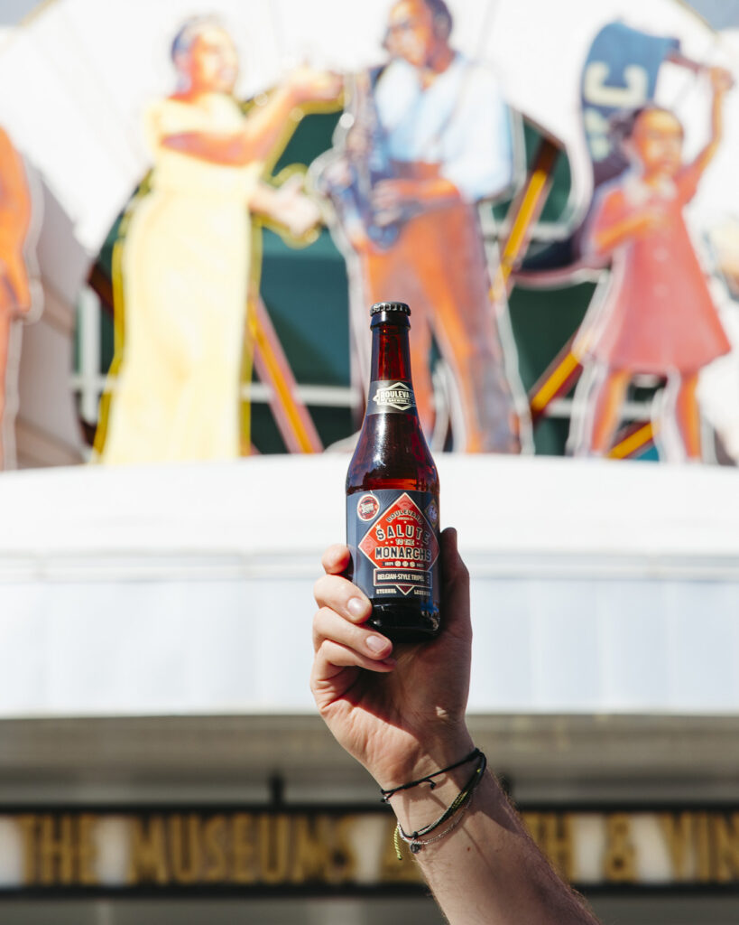 A person holds up a bottle of "Boldy Honest" root beer against the background of a colorful mural featuring musicians. The hand is slightly blurred while the bottle and its label are in sharp focus. The sign for "The Museum..." is partially visible at the bottom.