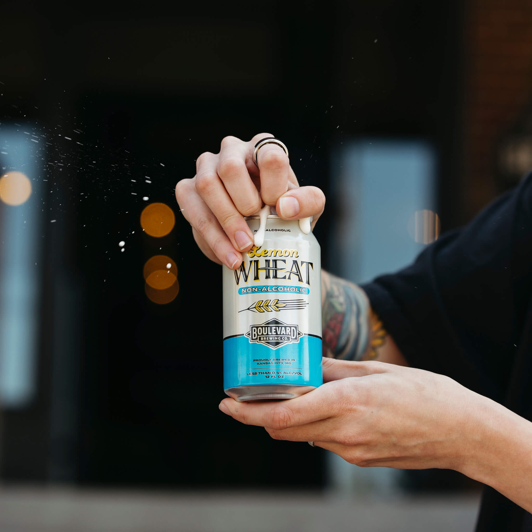 A person with tattoos on their arm is holding and opening a can of Boulevard Brewing Co.'s non-alcoholic wheat beer with lemon. Beer droplets are seen in the air as the can is being opened. The background is blurred, focusing on the can and the person's hands.