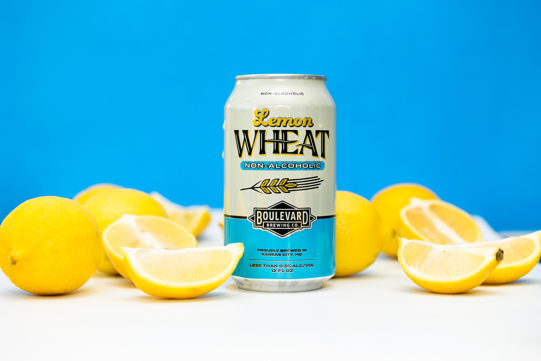 A can of Boulevard Brewing Co.'s Non-Alcoholic Lemon Wheat beer stands against a blue background. The can is surrounded by whole and halved lemons, emphasizing the lemon flavor of the beverage. The label is light blue and white with yellow accents.