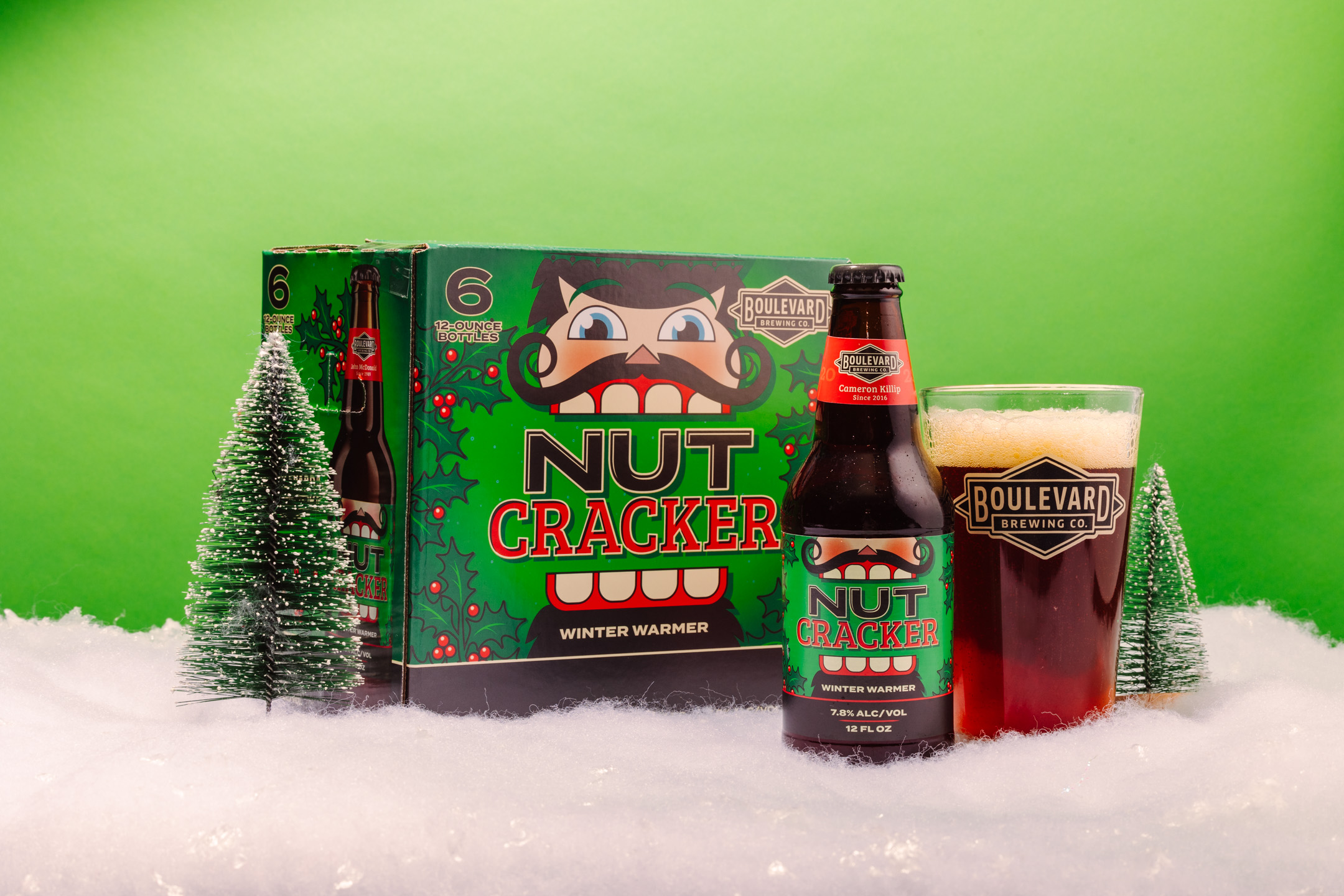 A bottle and a six-pack of Boulevard Brewing Co.'s Nutcracker Winter Warmer beer placed on artificial snow with a clear glass of beer next to it. Two small model pine trees are also in the scene, against a bright green background.