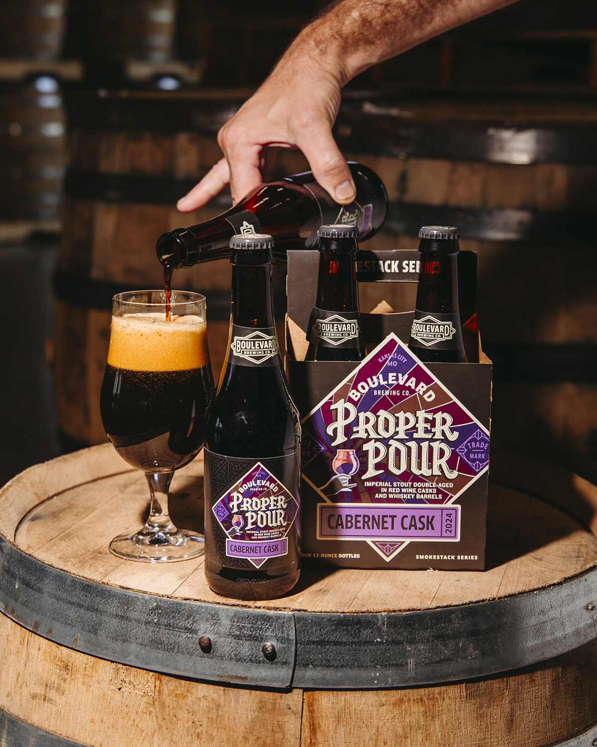 A hand pours Boulevard Proper Pour Cabernet Cask beer into a glass on a wooden barrel. A four-pack of the same beer and an additional bottle are placed beside the glass. The labels are purple and white. A rustic background with wooden barrels is visible.