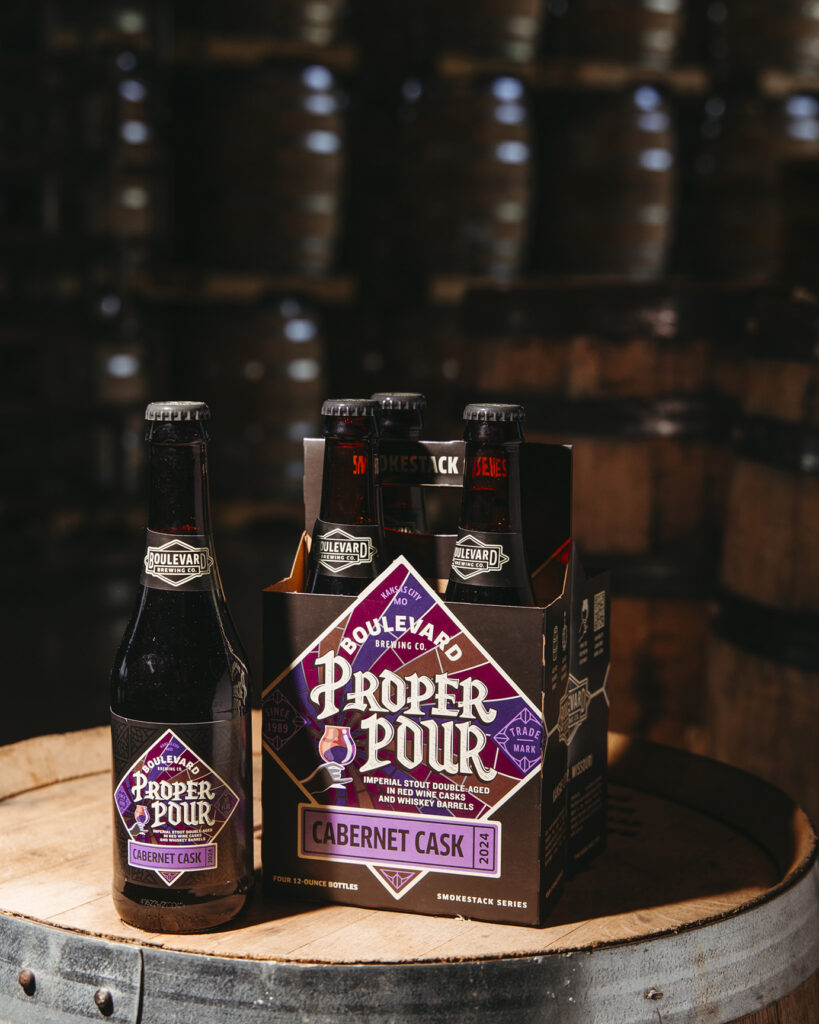 A four-pack of Boulevard Brewing Co.'s Proper Pour Cabernet Cask beer sits on a wooden barrel. One bottle is placed beside the pack. The scene is lit softly, highlighting the product against a backdrop of blurred wooden barrels.