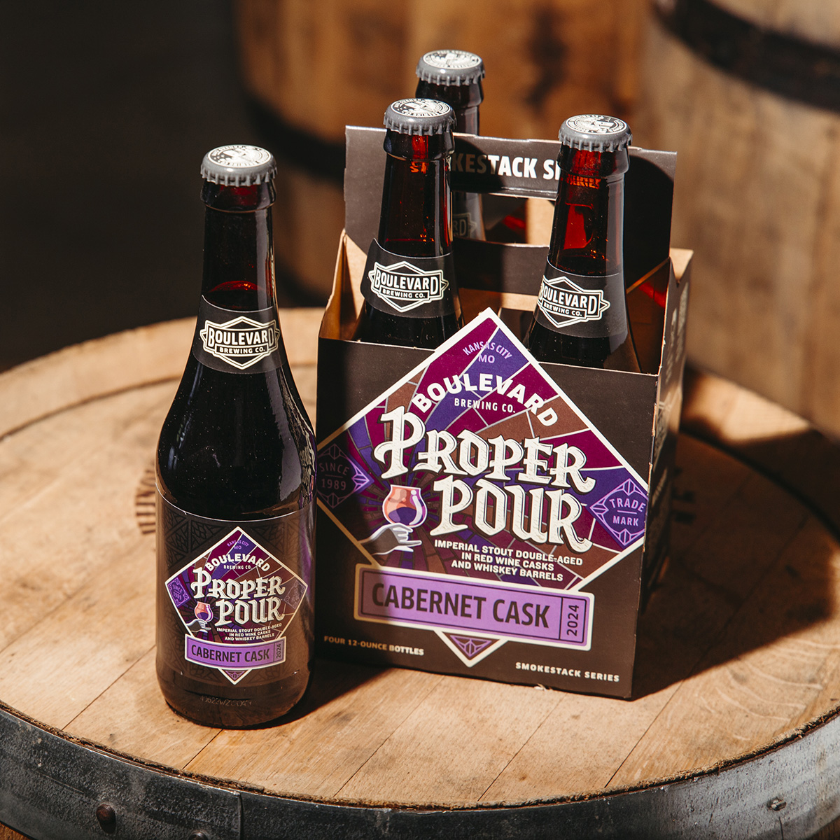 A bottle and a four-pack of Boulevard Brewing Co.'s Proper Pour Imperial Stout are displayed on top of a wooden barrel. The beer bottles have dark labels with purple accents and are labeled "Cabernet Cask" as part of the Smokestack Series.