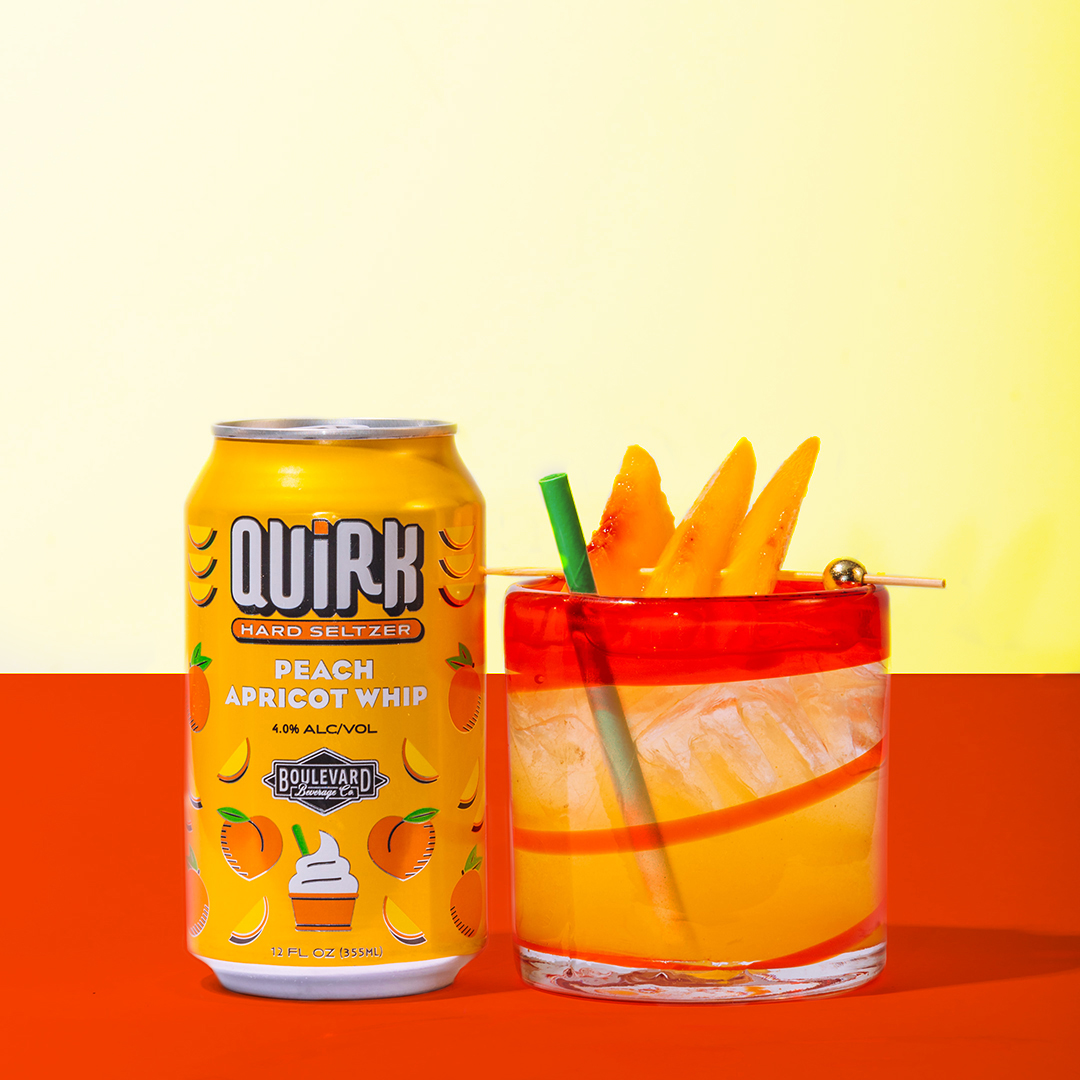 Image shows a can of Quirk Hard Seltzer Peach Apricot Whip next to a cocktail glass filled with ice and a peach-colored beverage. The glass is garnished with slices of peach or apricot on a pick and has a green straw. The background is a gradient of yellow and orange.