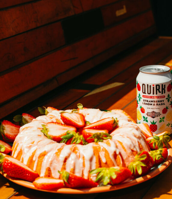 A bundt cake drizzled with glaze and garnished with fresh strawberries is placed on a wooden surface. Nearby, there is a can of Quirk Strawberry Lemon & Basil hard seltzer. The scene is warmly lit, creating a cozy and inviting atmosphere.