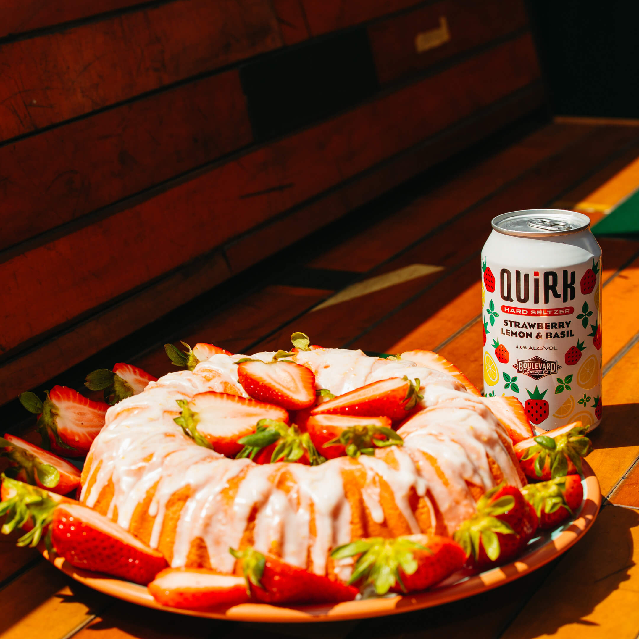 A bundt cake drizzled with glaze and garnished with fresh strawberries is placed on a wooden surface. Nearby, there is a can of Quirk Strawberry Lemon & Basil hard seltzer. The scene is warmly lit, creating a cozy and inviting atmosphere.