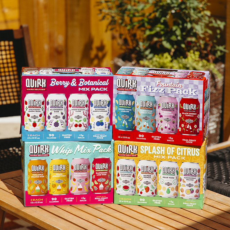 Four colorful Quirk Hard Seltzer variety packs are stacked outdoors on a sunlit table with a wooden fence in the background. Each pack displays different flavors: Berry & Botanical Mix Pack, Fountain Fizz Pack, Whip Mix Pack, and Splash of Citrus Mix Pack.