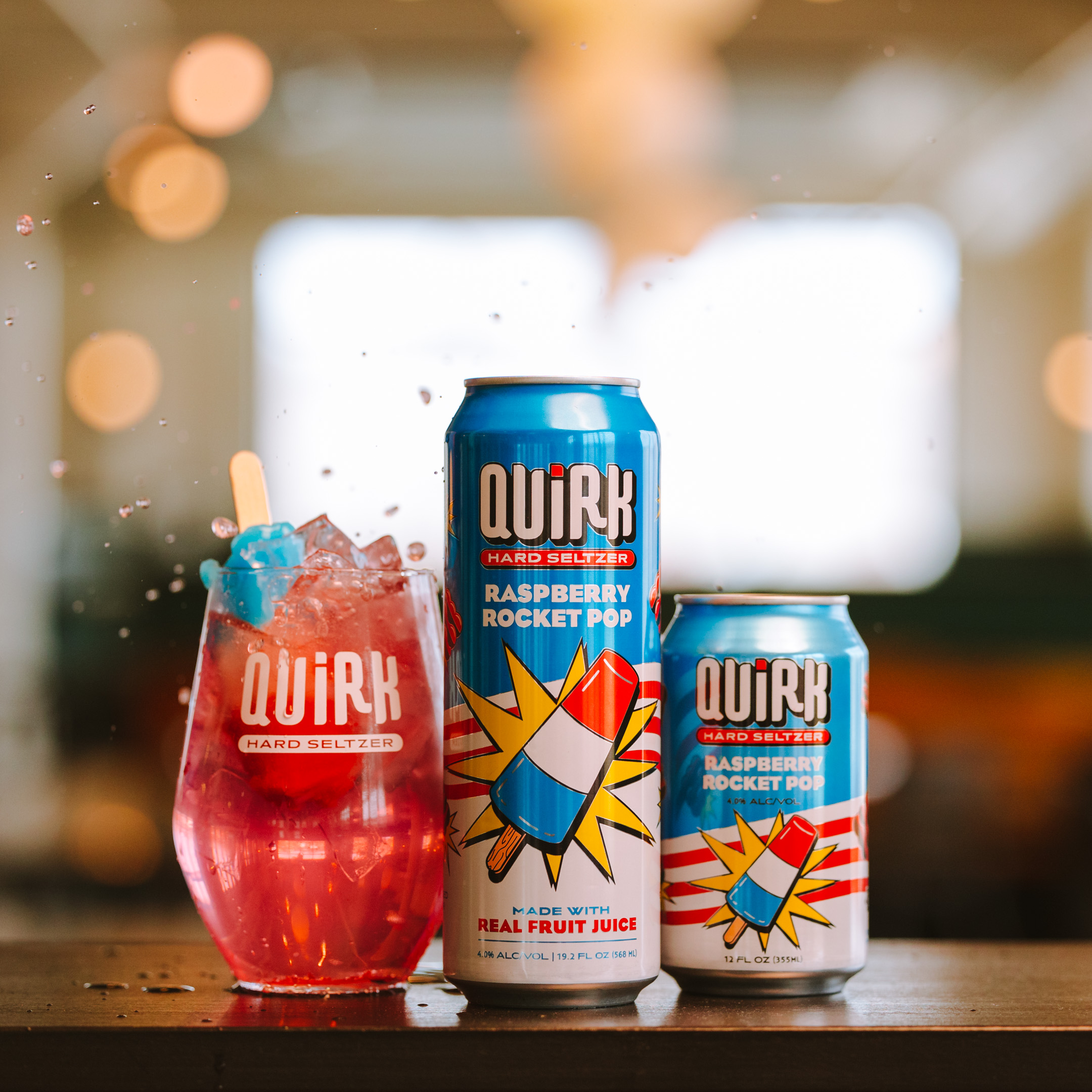 A can and a smaller can of Quirk Hard Seltzer Raspberry Rocket Pop flavor are placed on a table alongside a glass filled with the same drink, featuring a popsicle submerged inside. The beverage is spilling over the glass, creating a dynamic, splashy scene.