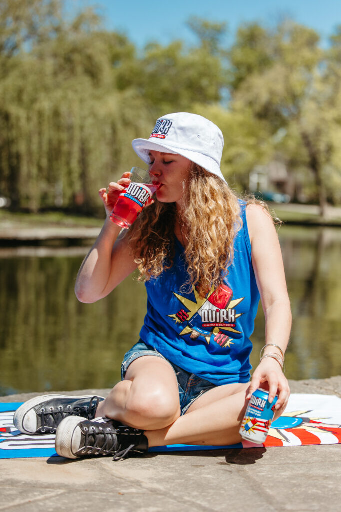 A young woman with long curly hair sits on a blanket outdoors by a lake, drinking a red beverage from a cup. She wears a blue tank top, a white bucket hat, denim shorts, and black sneakers. She holds a can in her other hand, which matches the branding on her shirt.