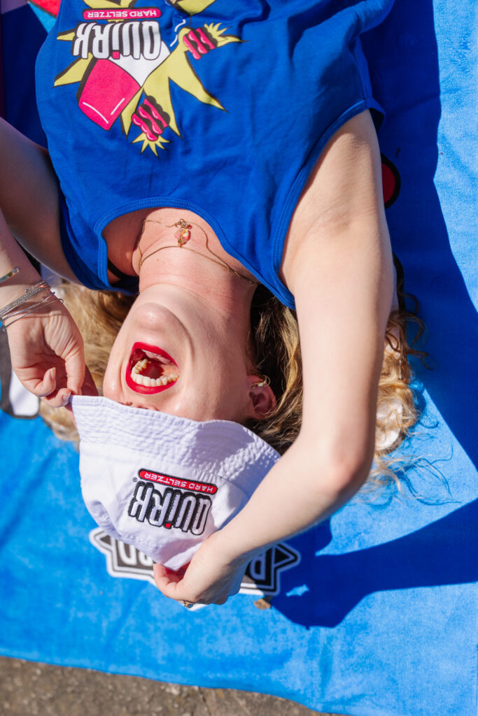 A person lies on a blue towel, wearing a blue tank top and holding a white bucket hat with the "Quirk Hard Seltzer" logo covering their face. They are laughing with their mouth open, showing visible excitement. Sunlight brightens the scene.