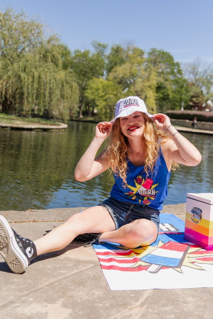 A person with long blonde hair, wearing a colorful tank top, denim shorts, and a white bucket hat, is sitting by a serene lake. They're smiling and adjusting their hat. Near them are a striped mat and a small box. The background shows lush green trees and a calm water body.