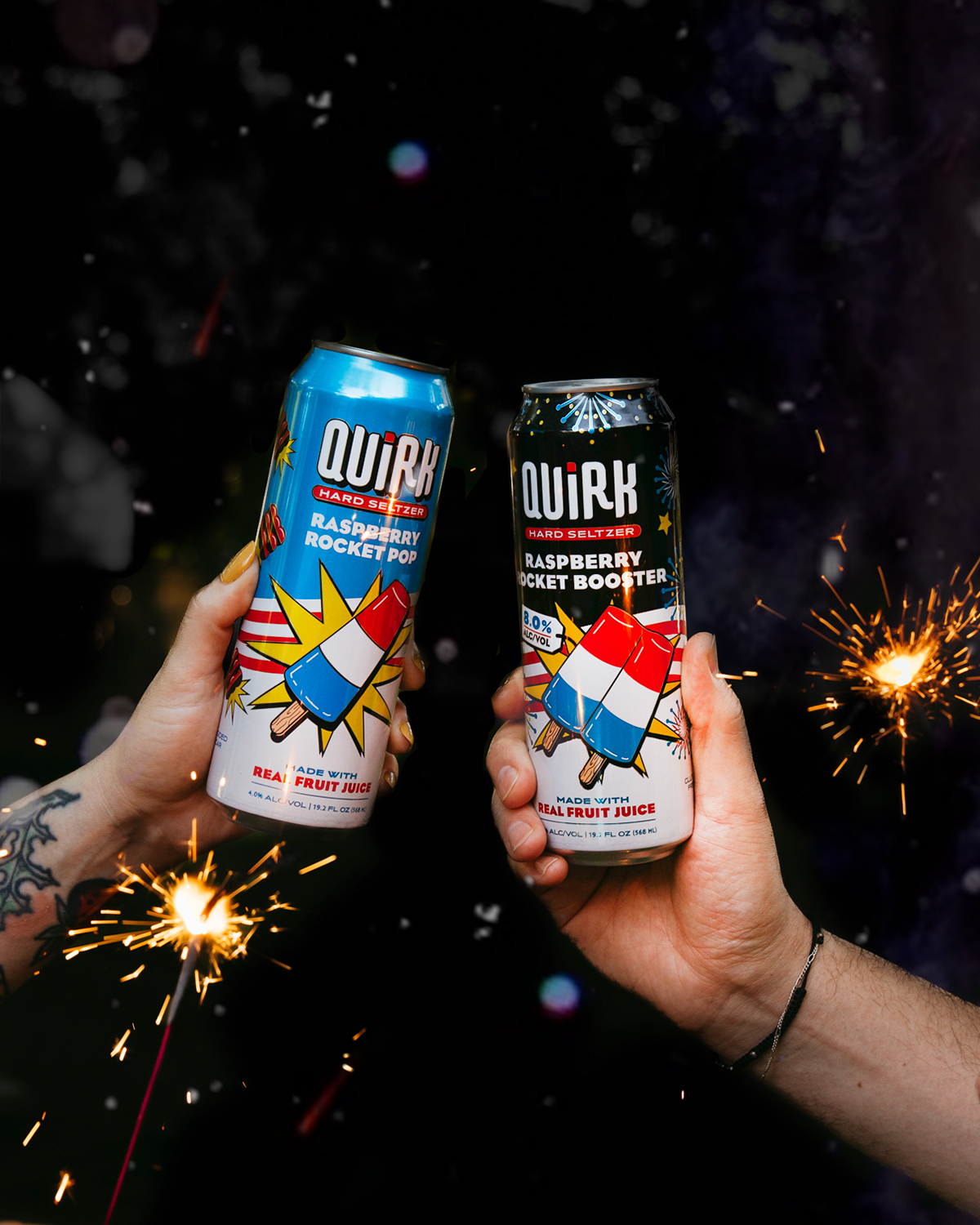 Two hands hold up cans of Quirk Hard Seltzer in front of a dark background with sparklers. The cans are labeled "Raspberry Rocket Pop" and "Raspberry Rocket Booster" and feature vibrant graphics of a rocket pop popsicle.