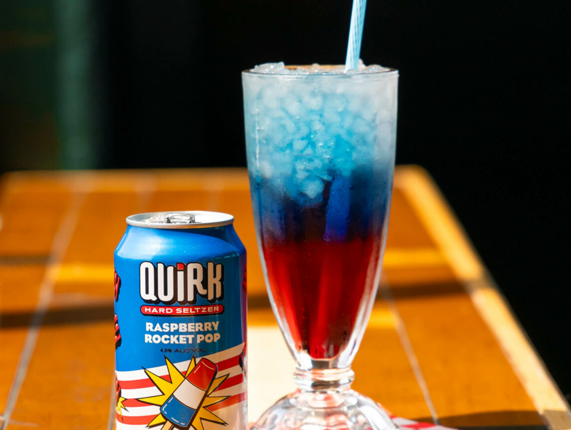 A can of Quirk Hard Seltzer labeled "Raspberry Rocket Pop" stands next to a tall glass of red and blue layered beverage with ice and a straw. The drink and can are set on a wooden surface with a checkered napkin underneath.