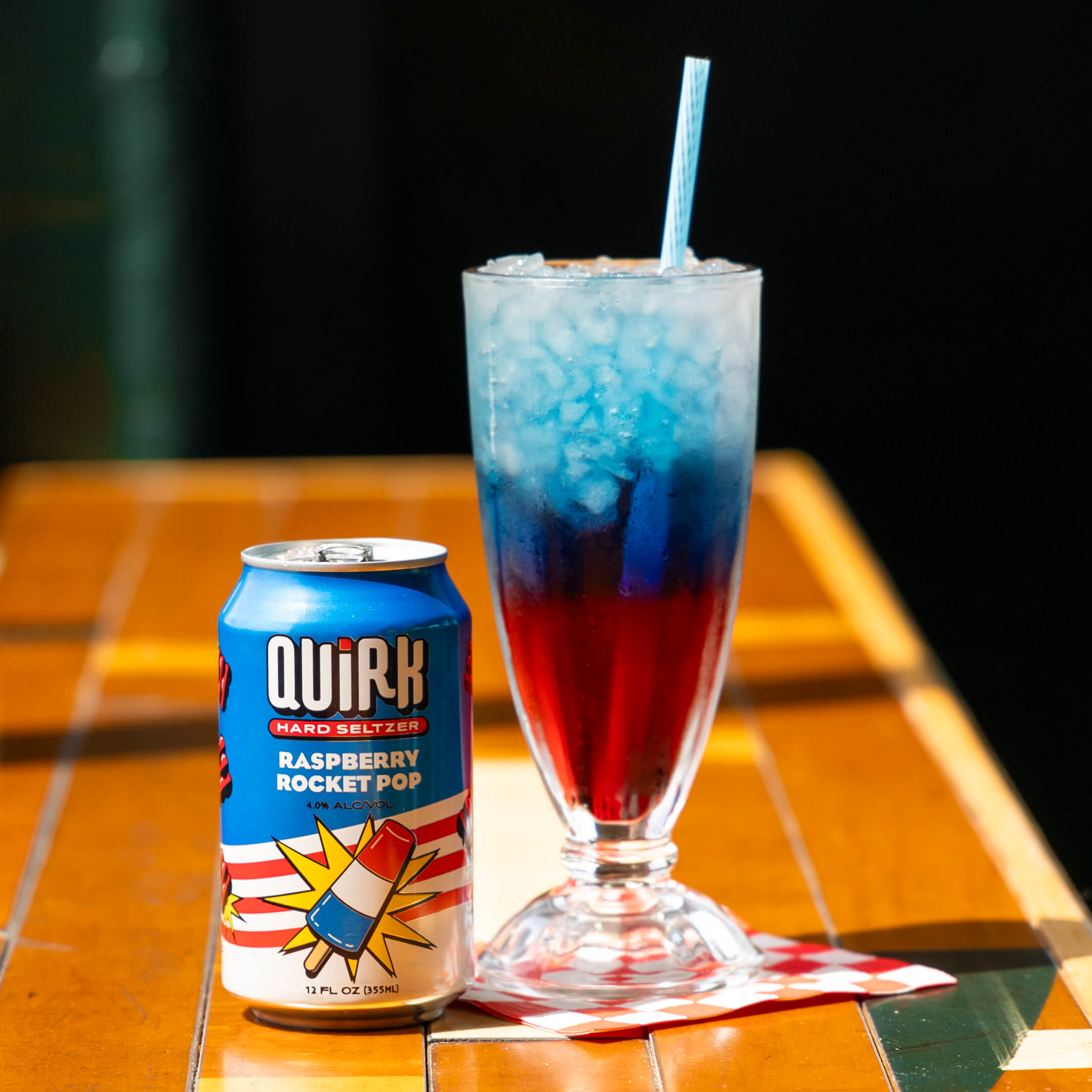 A can of Quirk Hard Seltzer labeled "Raspberry Rocket Pop" stands next to a tall glass of red and blue layered beverage with ice and a straw. The drink and can are set on a wooden surface with a checkered napkin underneath.