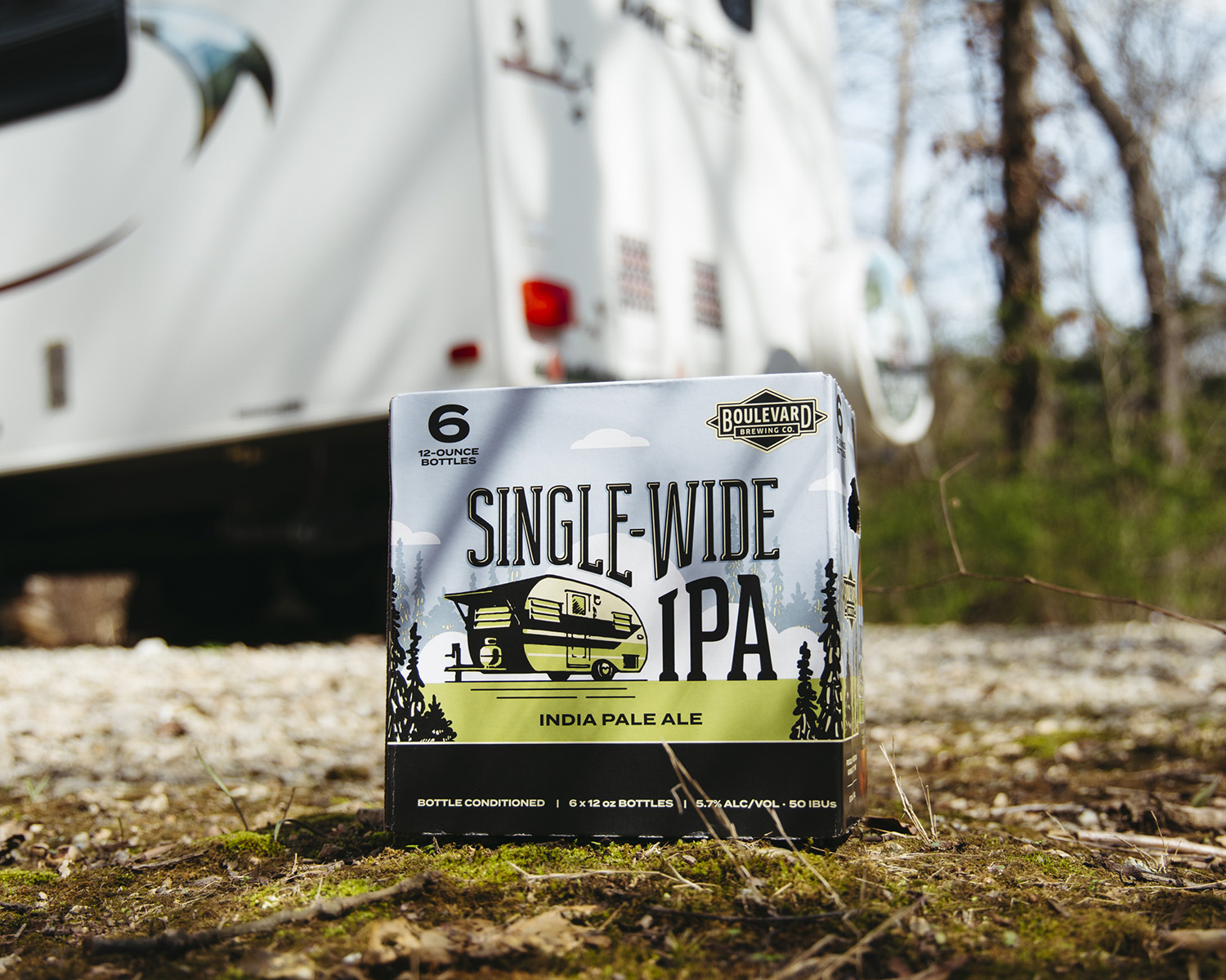 A six-pack of Boulevard Single-Wide IPA beer positioned outdoors in front of a camper. The packaging is green, black, and white, with a vintage trailer illustration. Sunlight filters through trees in the background, giving the scene a rustic and adventurous feel.