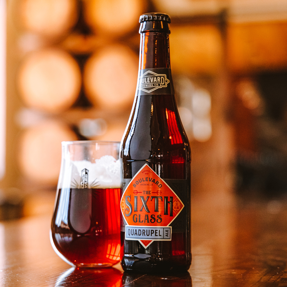 A bottle of Boulevard Brewing Co.'s The Sixth Glass Quadrupel Ale stands next to a full glass of the same ale. The rich amber beer has a foamy head, and the bottle label features a red and orange geometric design with the beer's name prominently displayed.