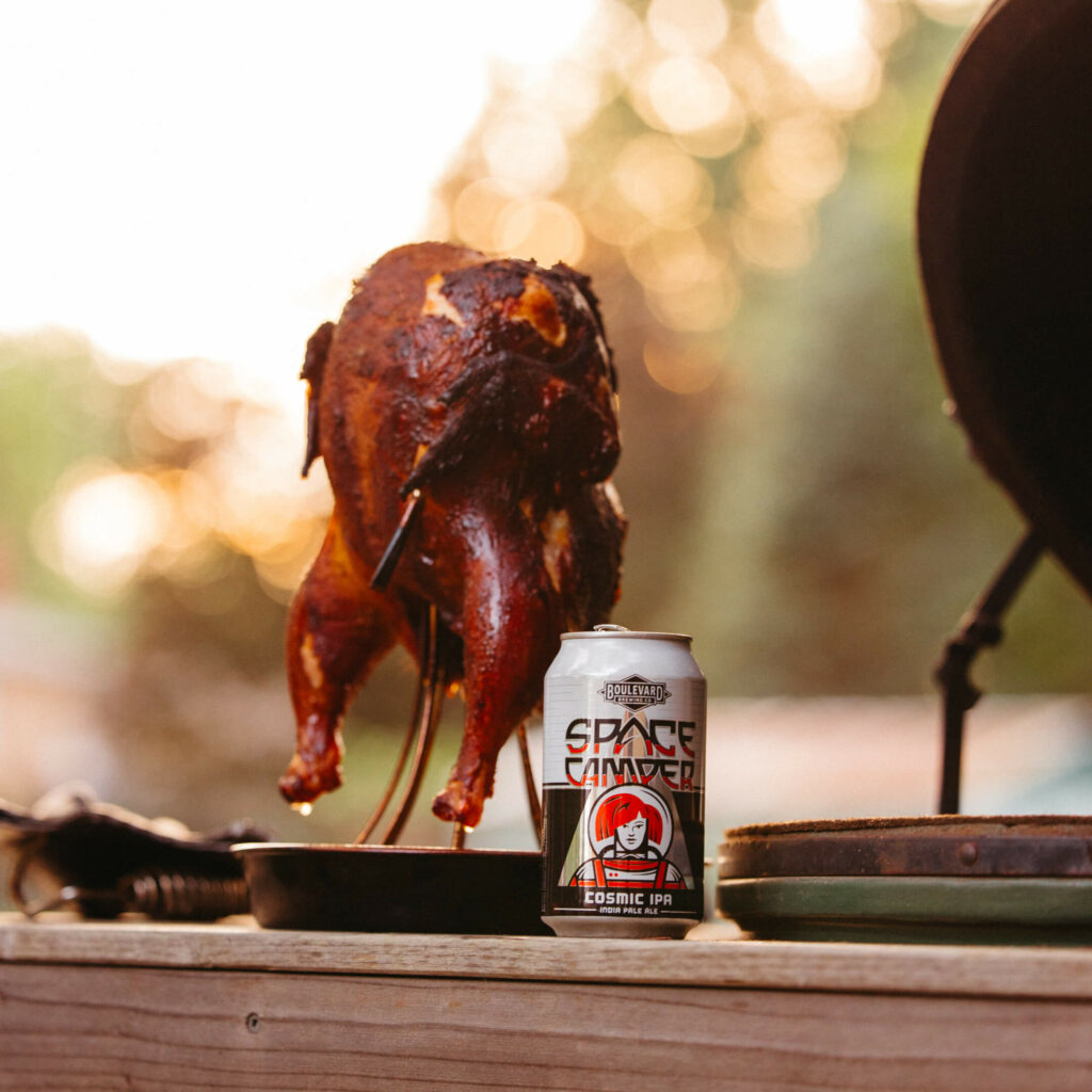 A roasted chicken on a grill with a can of Space Camper Cosmic IPA beer placed in front of it. The sun sets in the background, casting a warm glow over the scene. The beer can features a colorful, astronaut-themed design.