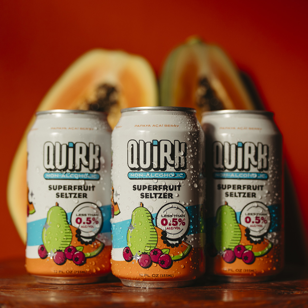 Three cans of Quirk Non-Alcoholic Superfruit Seltzer are displayed against a backdrop of halved papayas. The cans feature vibrant labels with images of mangoes, papayas, and acai berries, highlighting the seltzer's 0.5% alcohol content.