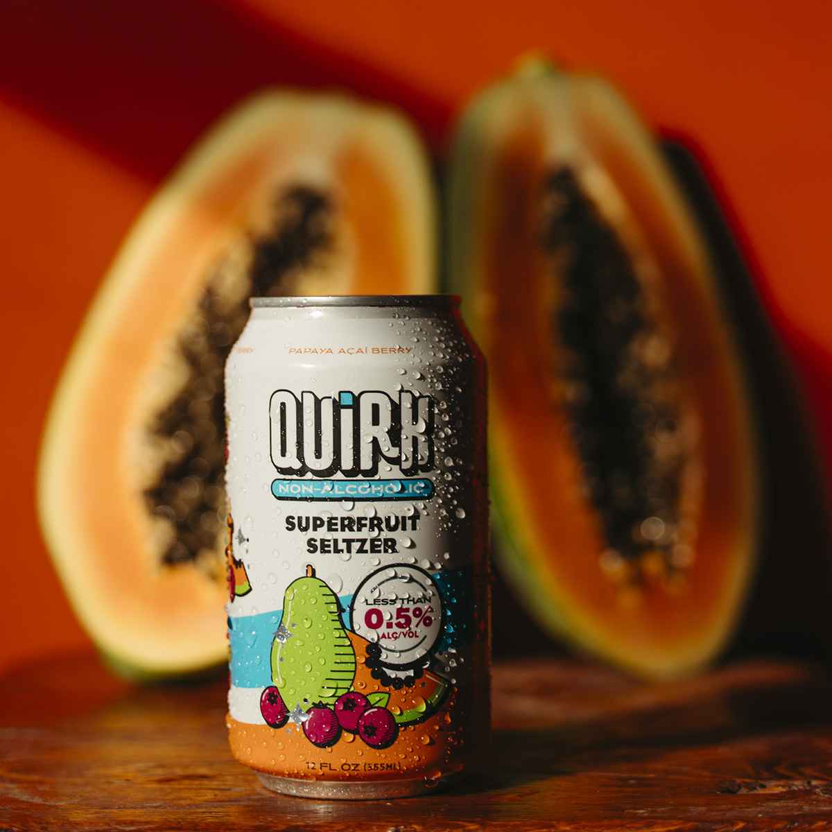 A can of Quirk Non-Alcoholic Superfruit Seltzer is displayed in front of two halves of a papaya. The label on the can features colorful fruit illustrations, including papaya and acai berries, with a bold 0.5% ABV marker and an orange background.