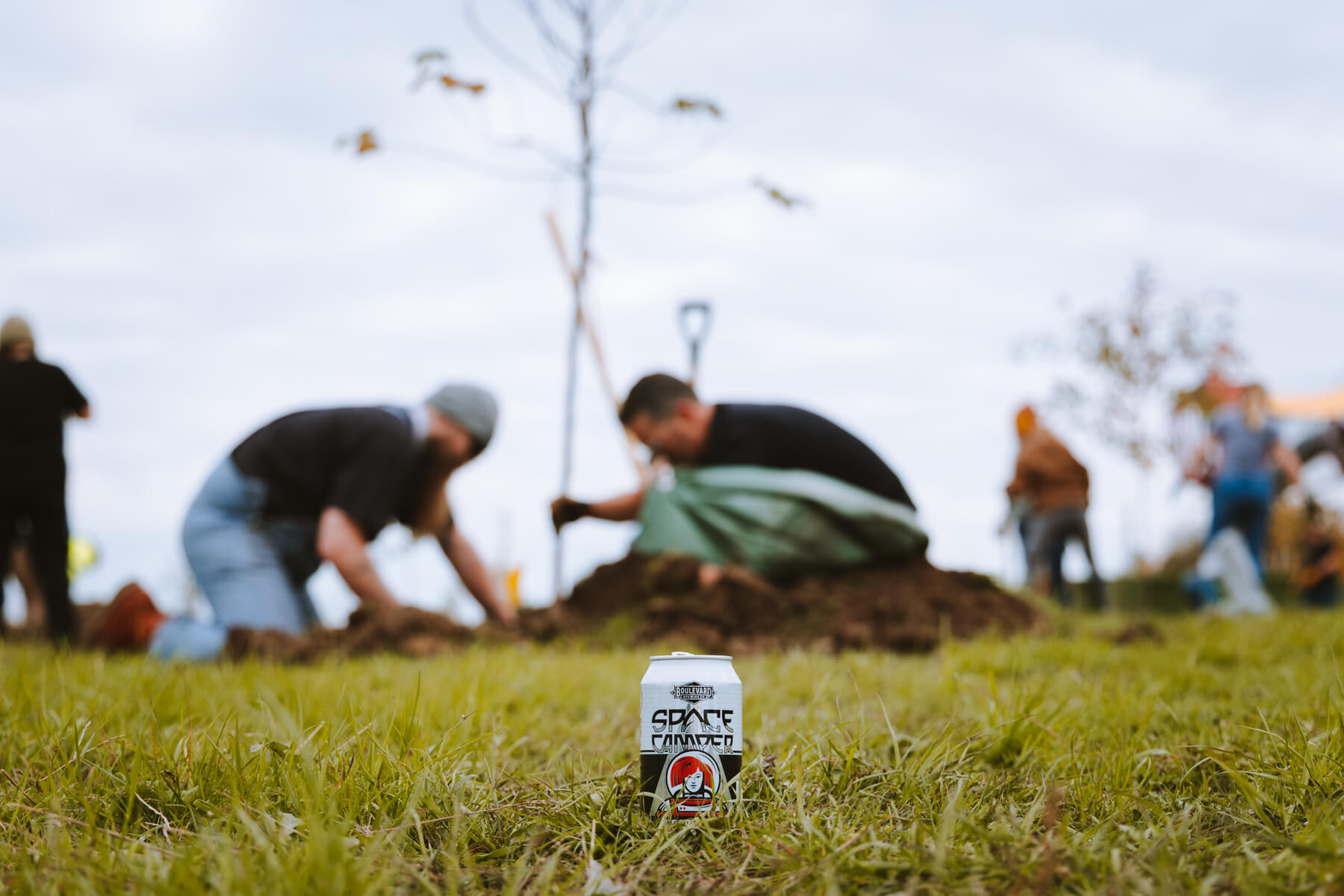 A can of Space Invader beer sits on the grass in the foreground. In the background, two people are planting a tree, with shovels and other tools scattered around. The sky is overcast, and other people can be seen working further in the distance.