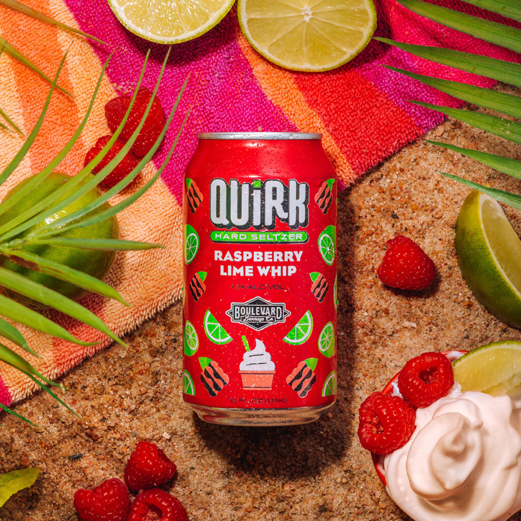 A can of Quirk Hard Seltzer, Raspberry Lime Whip flavor, is placed on sandy ground amidst fresh raspberries, lime slices, a tropical towel, and some whipped cream. Palm leaves partially frame the scene, creating a vibrant and summery atmosphere.