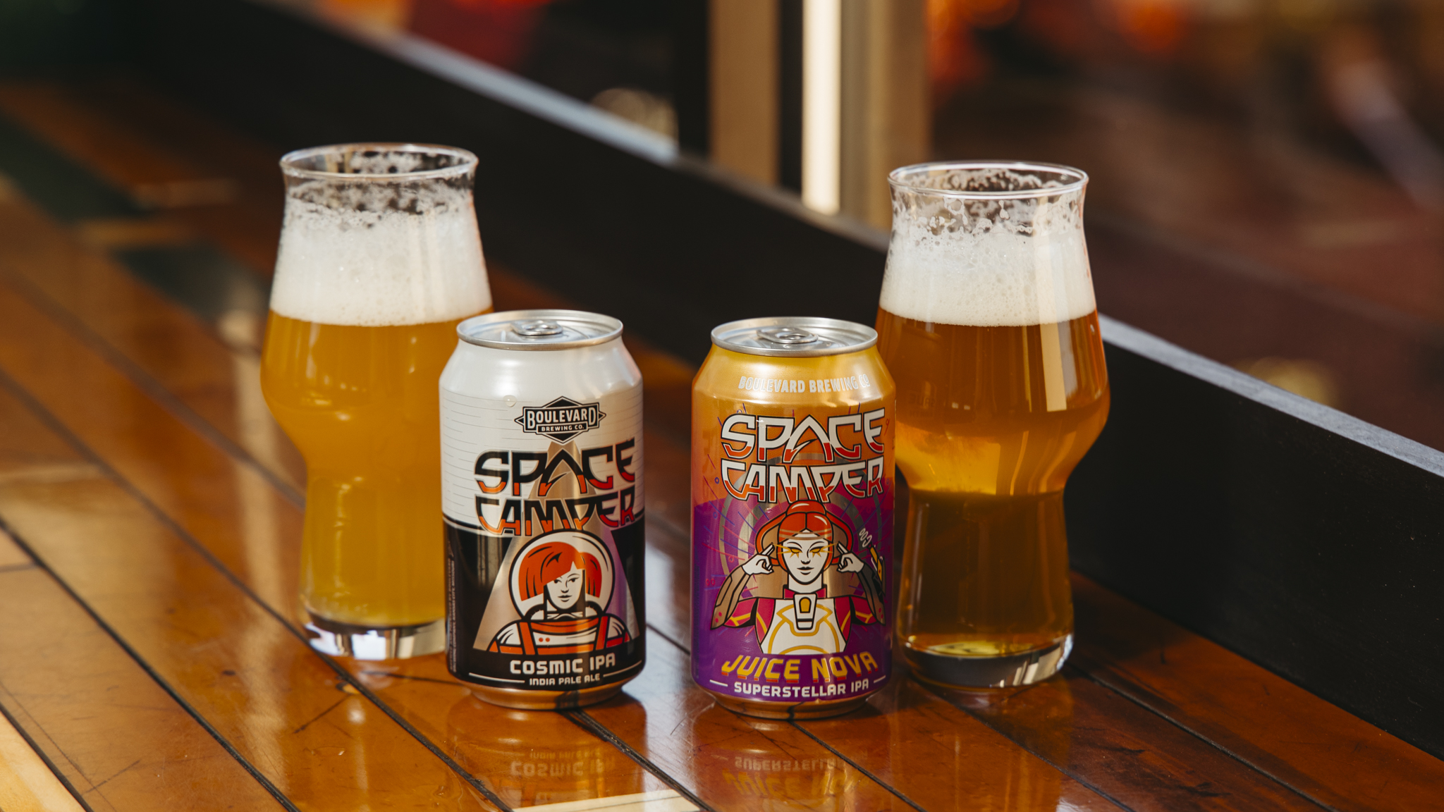 Two cans of Space Camper beer, Cosmic IPA and Space Camper Juice Nova, are on a wooden table, each paired with a filled glass. The cans boast vibrant designs featuring astronauts.