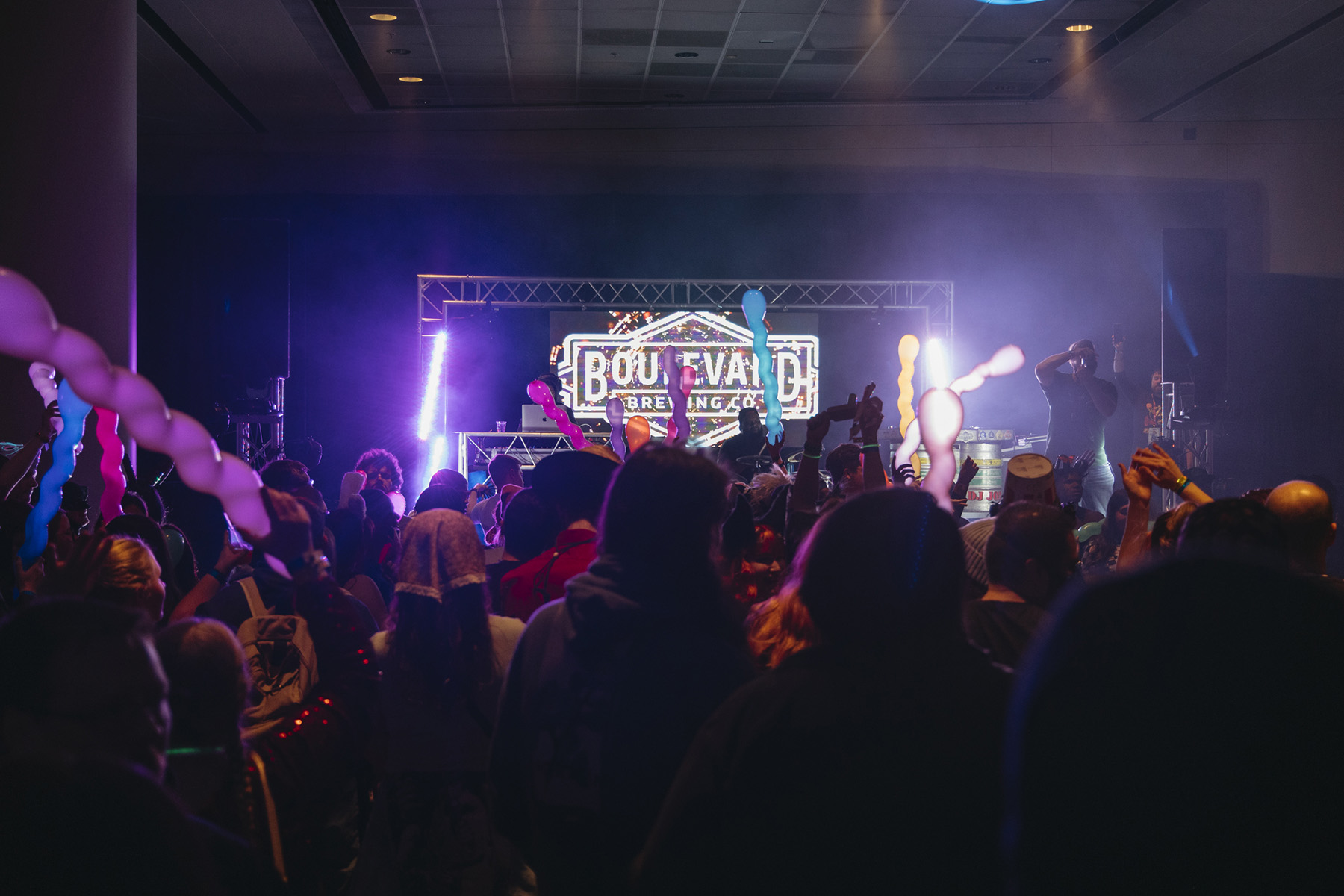 A lively concert scene buzzes with excitement as a crowd enthusiastically watches a performer on stage. A bright marquee sign boasting "BOULEVARD" lights up in the background. The audience, clutching glowing balloons, adds to the festive atmosphere under dim, colorful lighting all about them.