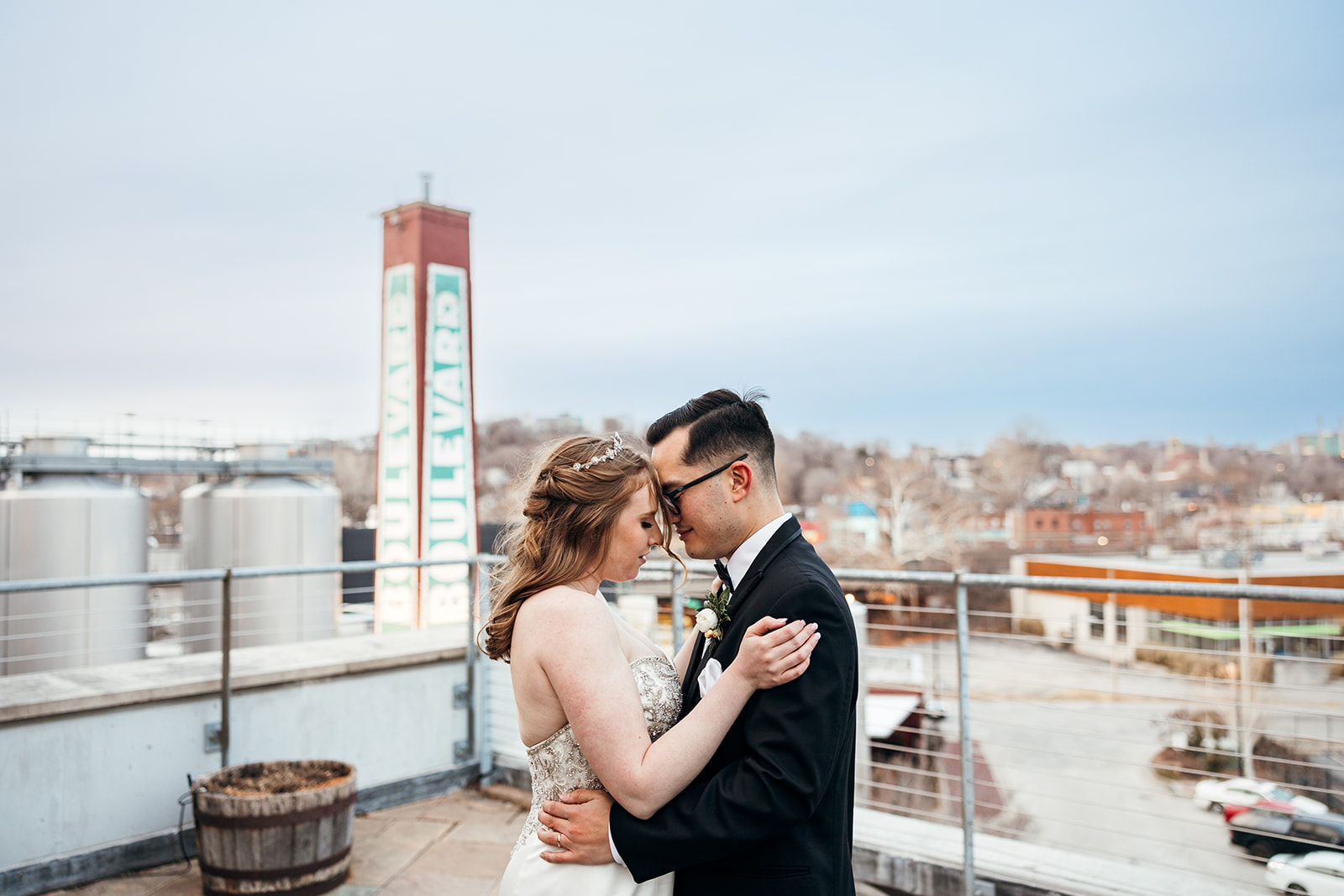 A bride and groom embrace on a rooftop, with an urban landscape in the background. A tall sign reading "Boulevard" is visible. The sky is overcast, adding a soft light to the scene.